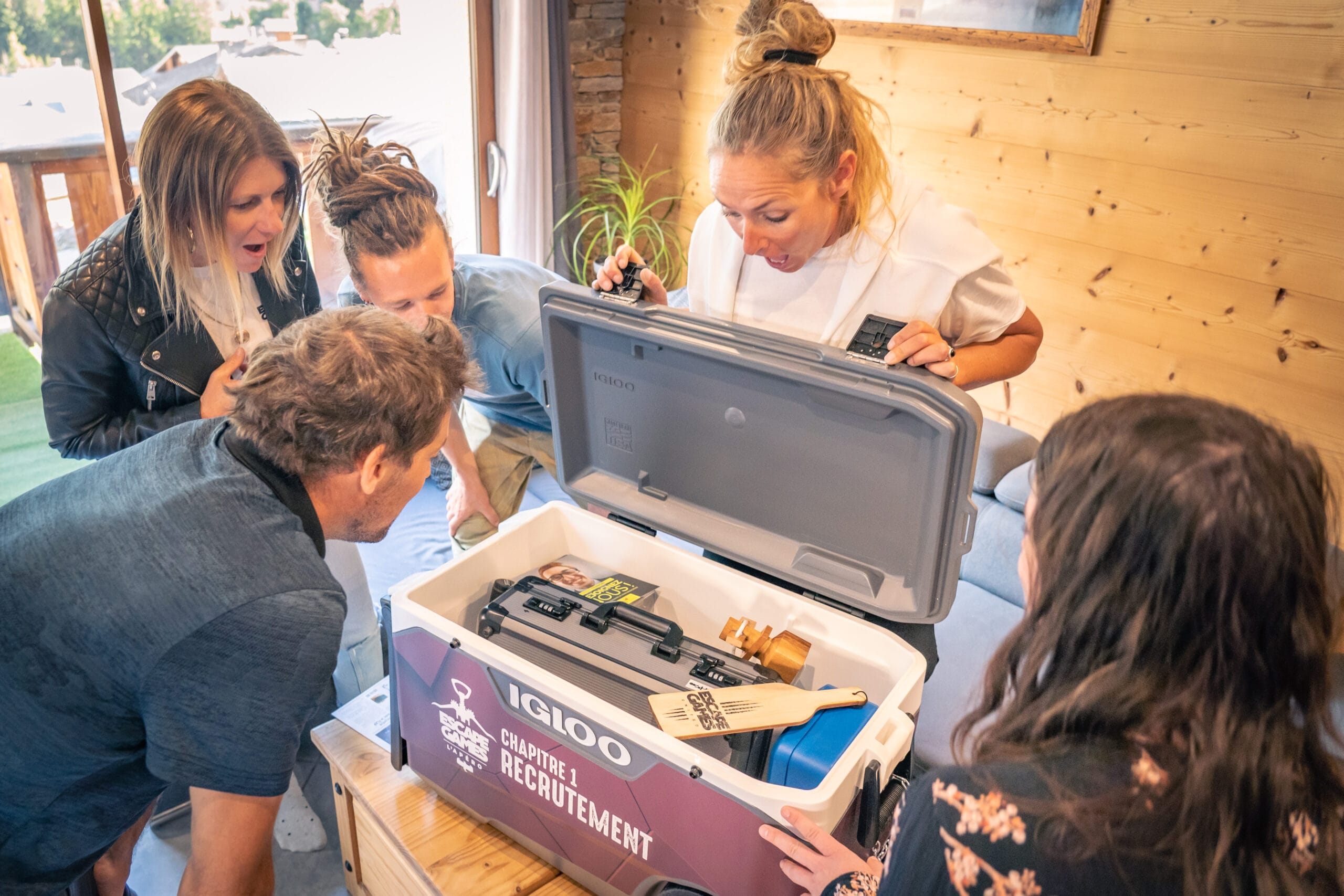 Un groupe d'amis exprime leur surprise et leur joie autour d'une glacière ouverte lors d'un escape game apéro. La glacière, utilisée comme coffre à secrets, contient des boissons et des snacks pour l'apéro, révélés seulement après avoir résolu des énigmes.